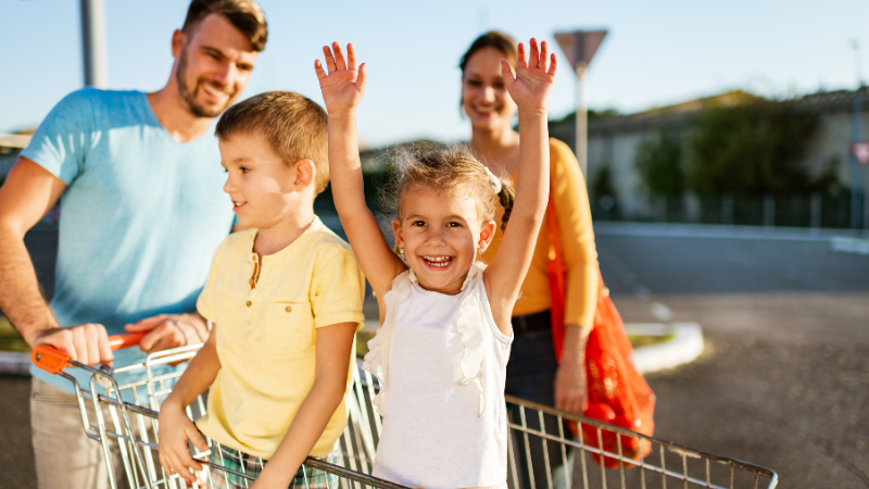 Two adults and two children stand outdoors with a shopping cart; one child sits inside the cart with arms raised.