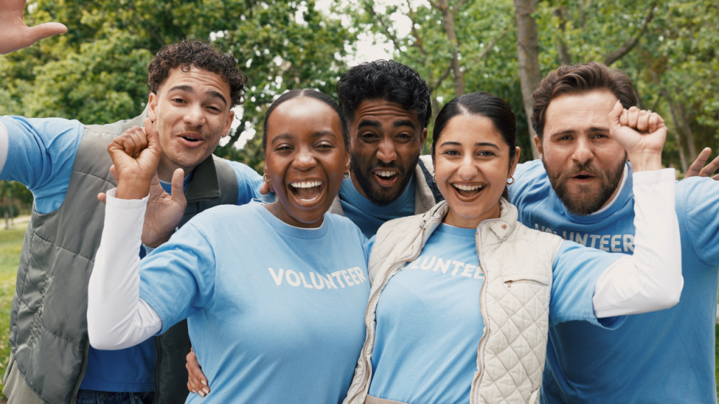 Five smiling volunteers in matching light-blue “VOLUNTEER” t-shirts huddle together outdoors under green trees, cheering with raised fists and big smiles.