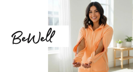 Person wearing a peach zip‑up jacket with slit sleeves, standing indoors near a window, with the text “Be Well” on the left.